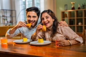 The Perfect Mother’s Day Gift: Swad Indian Kitchen. Indian Cuisine is the Perfect Choice for Mom. An Indian elderly mother and her young son are enjoying a delicious breakfast of dhokla, samosas, and fresh orange juice at a modern dining table, sharing joyful moments together.